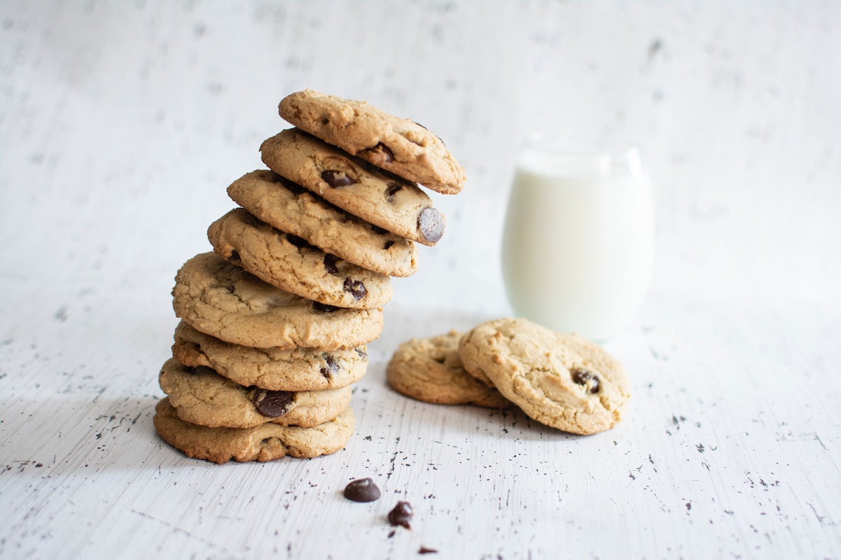 Leaning stack of chocolate chip cookies, next to a glass of milk, on a white table.