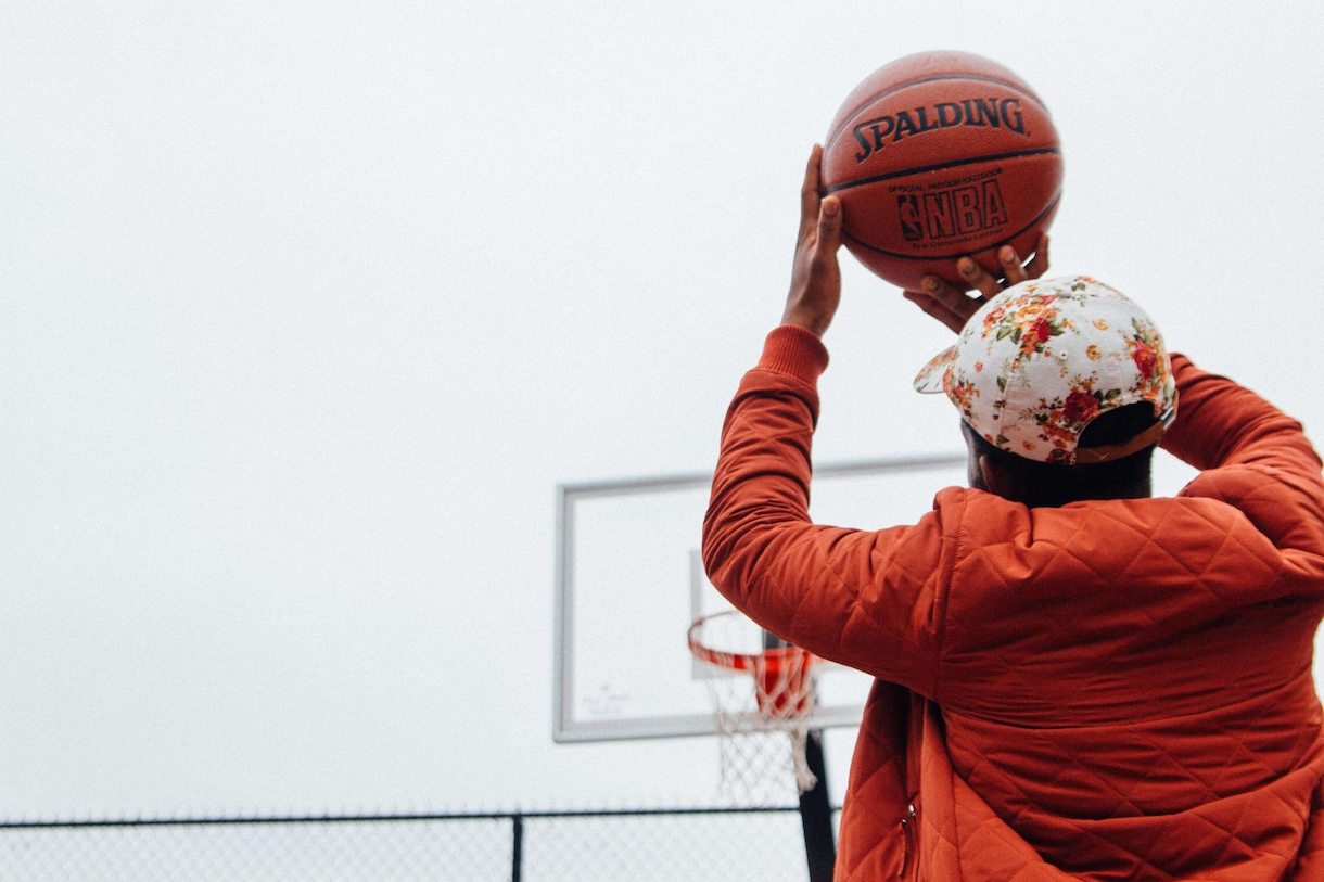 Person in orange jacket with flowered cap holding basketball above head about to shoot into hoop.